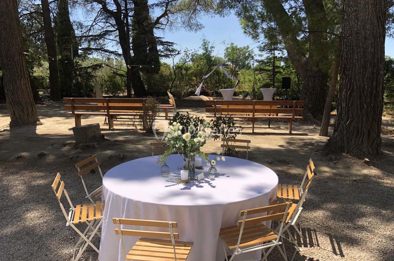 Table ronde décorée avec des fleurs et des chaises en bois, installée en extérieur sous des arbres, avec des bancs en…