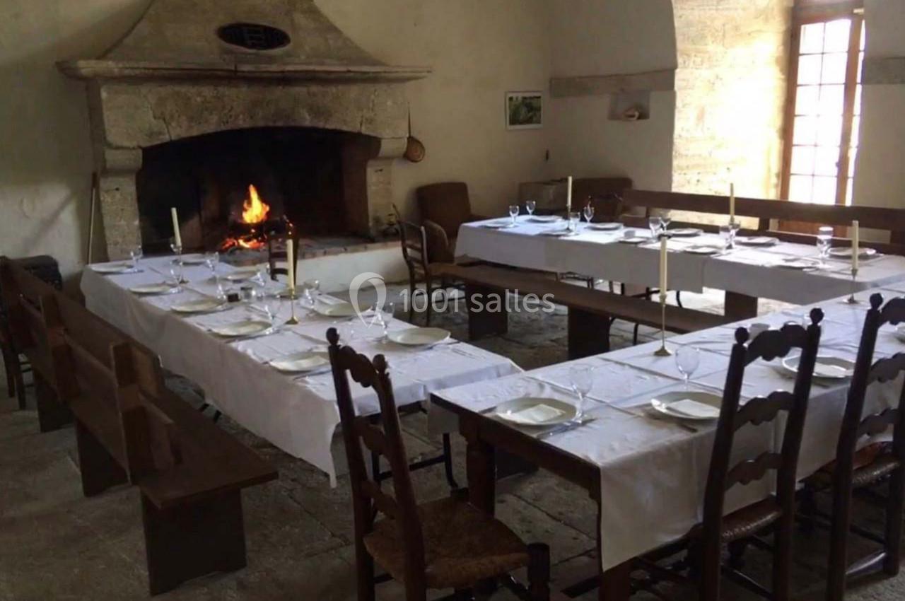 Salle à manger rustique avec tables dressées, nappes blanches, cheminée allumée et mobilier en bois.
