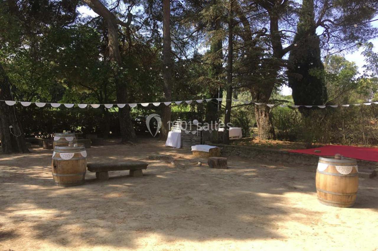 Espace extérieur ombragé avec guirlandes, tonneaux en bois, bancs rustiques et table blanche décorée.