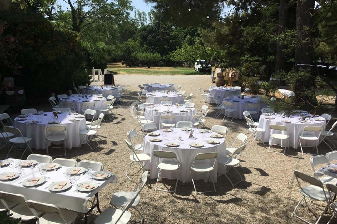 Tables rondes dressées avec nappes blanches et chaises pliantes dans un espace extérieur ombragé.