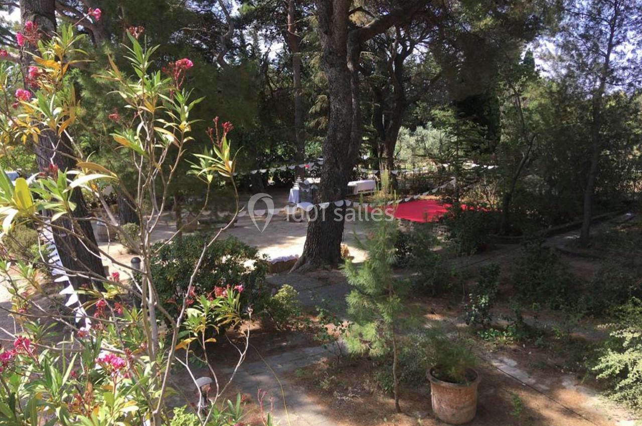 Jardin ombragé avec des arbres, des plantes fleuries, des guirlandes et des tables dressées en arrière-plan.