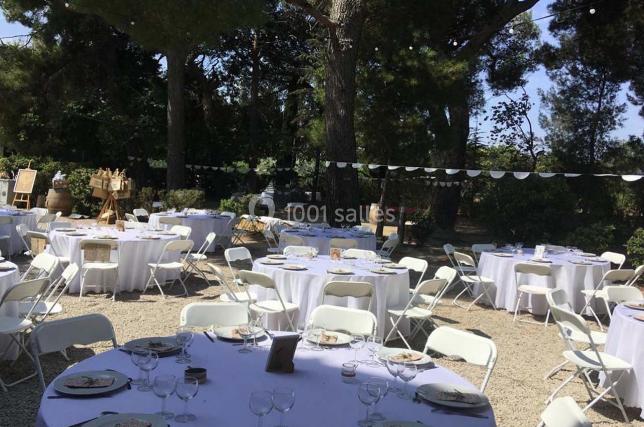 Tables rondes dressées avec nappes blanches et chaises pliantes dans un jardin ombragé, décoré pour un événement en plein…