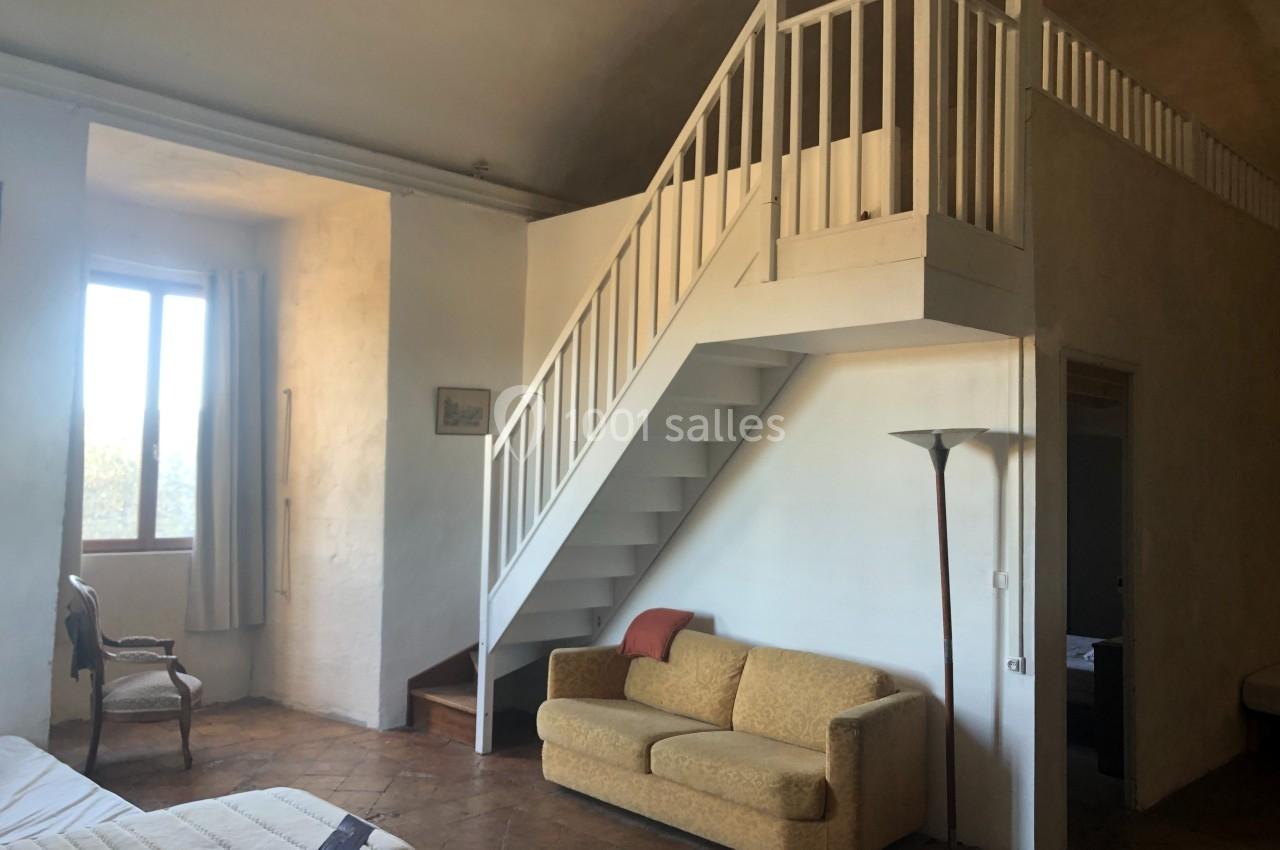 Intérieur d'une pièce avec un canapé, un escalier menant à une mezzanine et une fenêtre laissant entrer la lumière.
