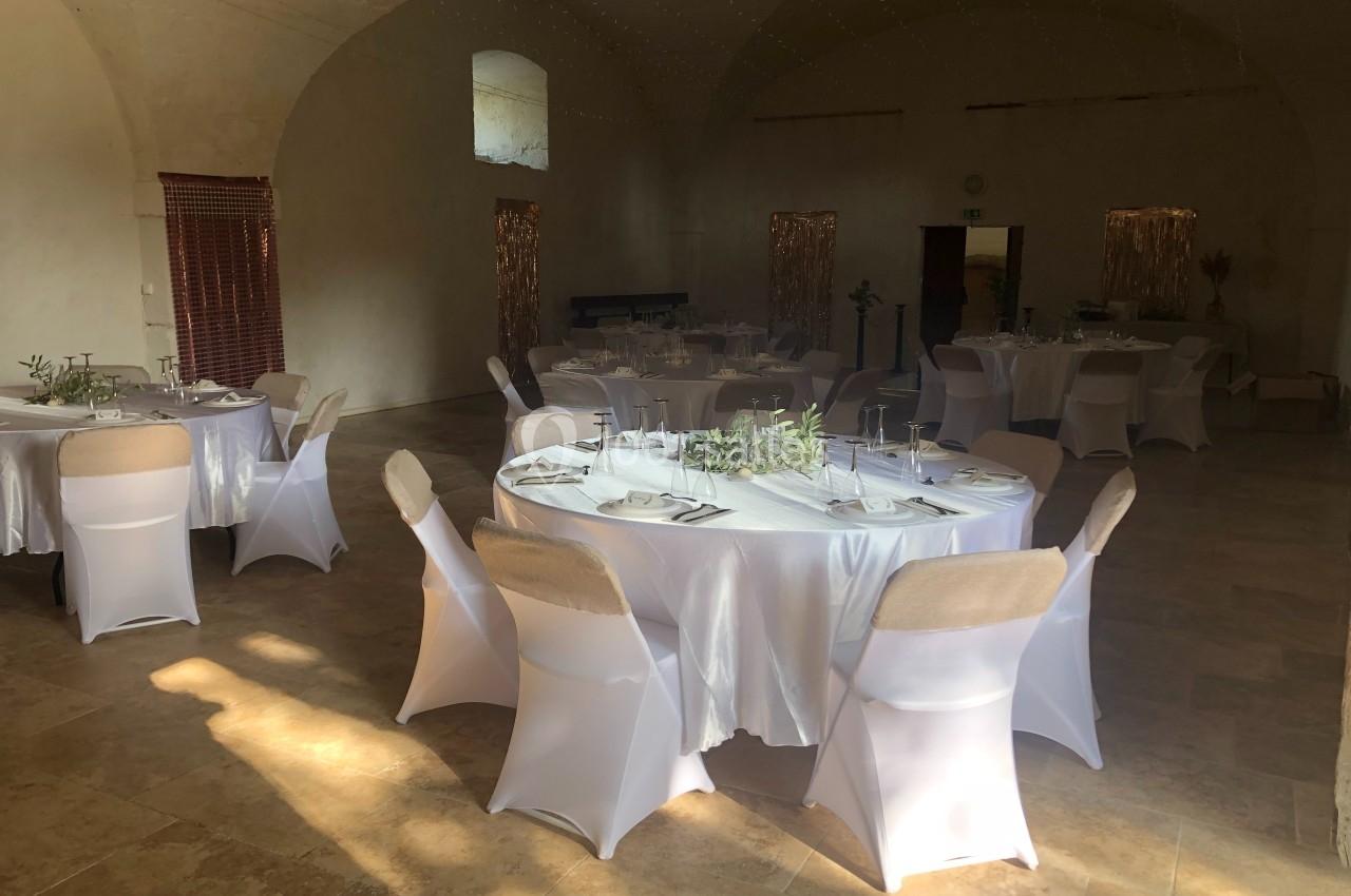 Salle de réception lumineuse avec tables rondes dressées, nappes blanches et chaises houssées, décor sobre et élégant.