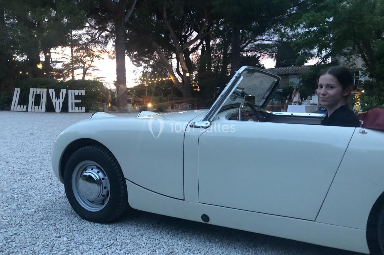 Une femme assise dans une voiture décapotable blanche, stationnée sur un chemin gravillonné, avec des arbres en arrière-plan.