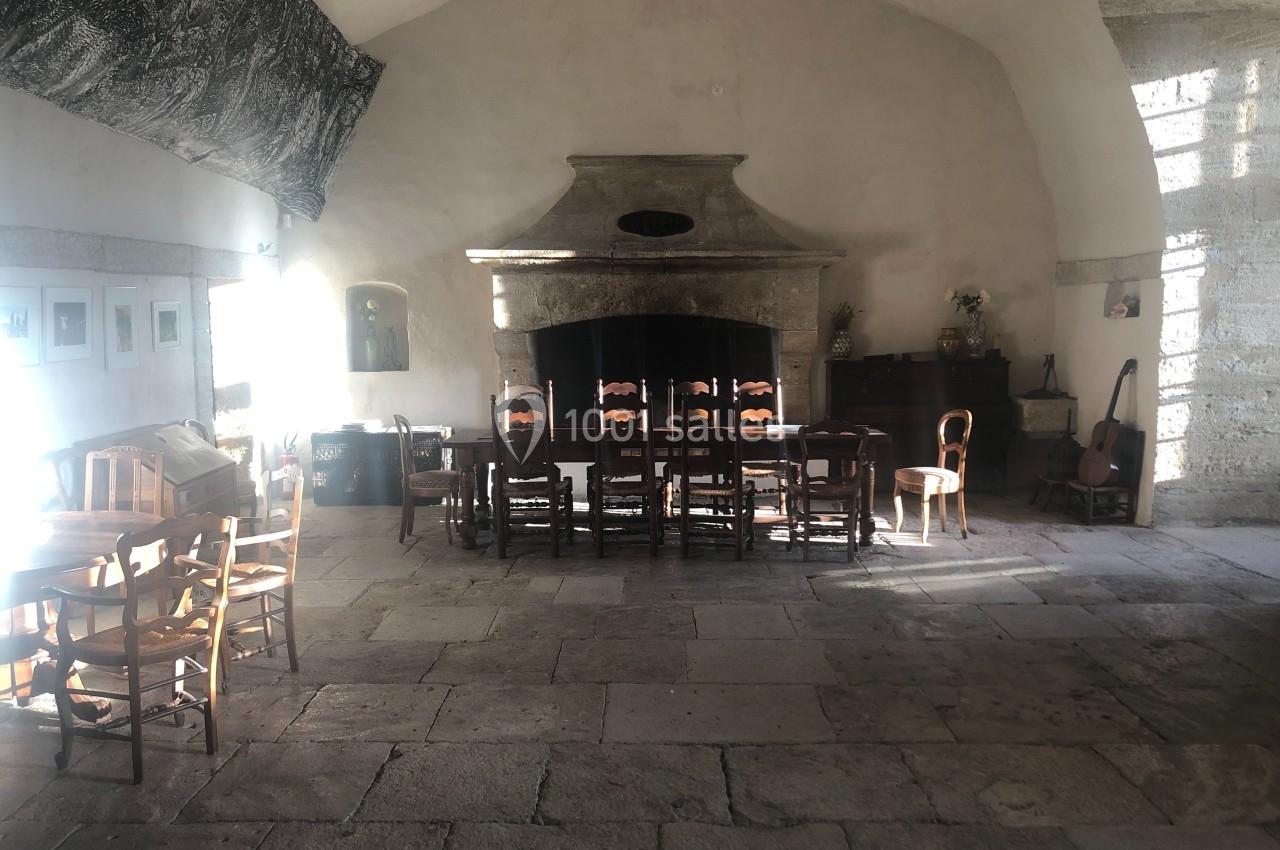 Grande pièce ancienne avec cheminée en pierre, table entourée de chaises en bois et lumière naturelle entrant par une…
