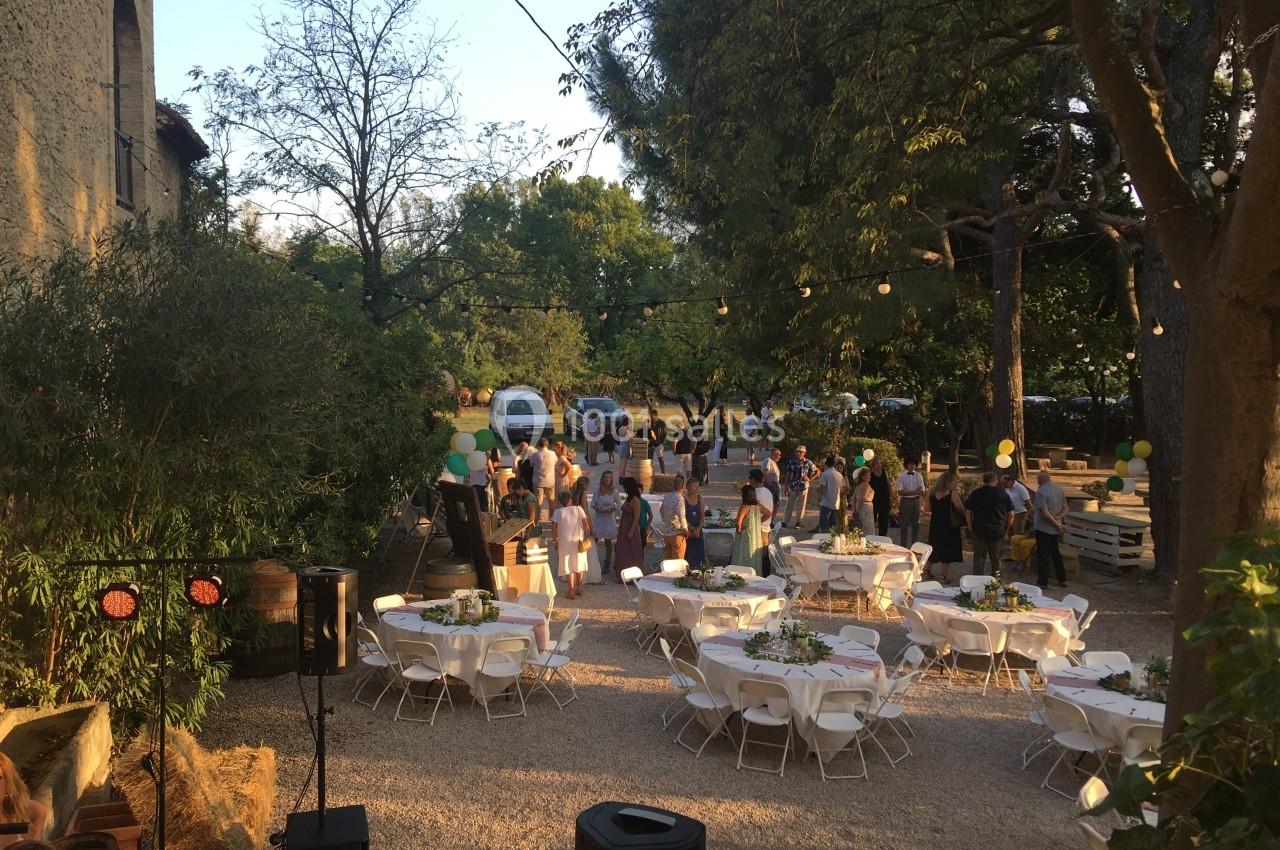 Groupe de personnes rassemblées dans une cour extérieure décorée, avec des tables rondes dressées et des guirlandes…