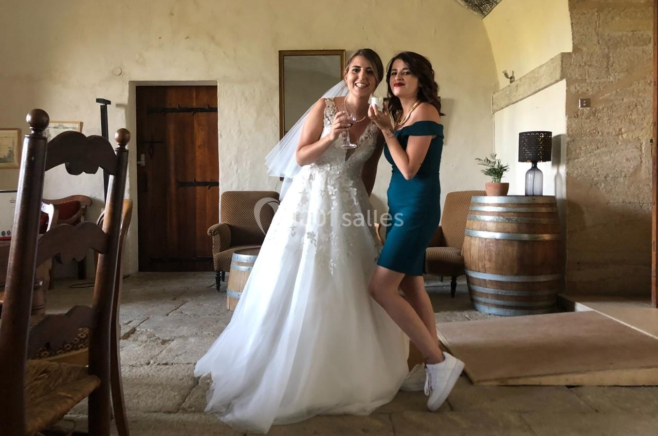 Deux femmes souriantes, l'une en robe de mariée et l'autre en robe verte, posent dans une pièce rustique.