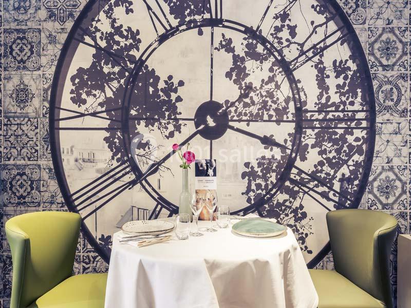 Table dressée pour deux avec vaisselle et fleurs, devant un mur décoré d'une grande horloge et de motifs carrelés.