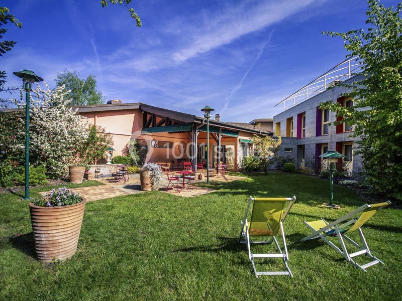 Jardin verdoyant avec chaises longues, terrasse aménagée et bâtiments colorés sous un ciel bleu dégagé.