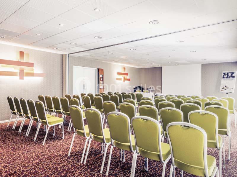 Salle de conférence lumineuse avec des rangées de chaises vertes alignées face à un écran de projection.