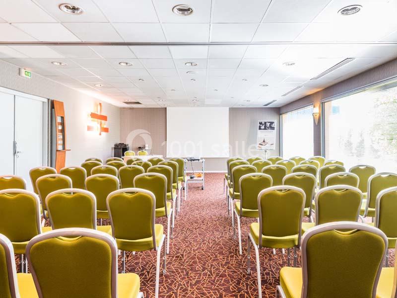 Salle de conférence lumineuse avec des rangées de chaises vertes alignées face à un écran de projection blanc.