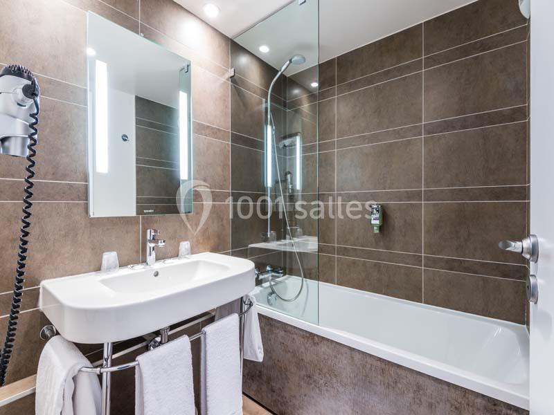 Salle de bain moderne avec lavabo, miroir éclairé, baignoire avec paroi vitrée et murs en carrelage marron.