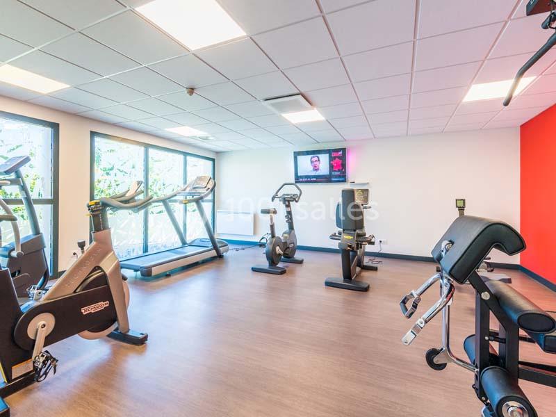 Salle de sport lumineuse avec tapis de course, vélos d'exercice, équipements de musculation et écran mural.