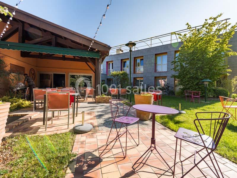 Cour extérieure ensoleillée avec tables et chaises colorées, entourée de bâtiments et de végétation.