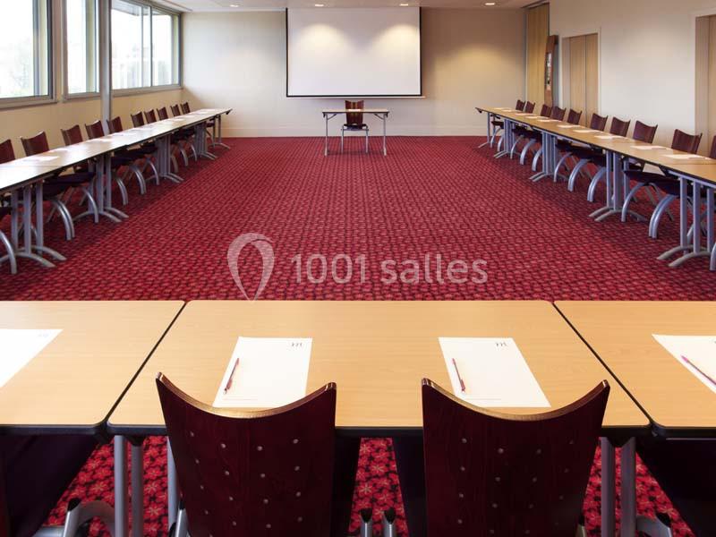 Salle de conférence avec tables disposées en U, chaises rouges et écran de projection au fond.