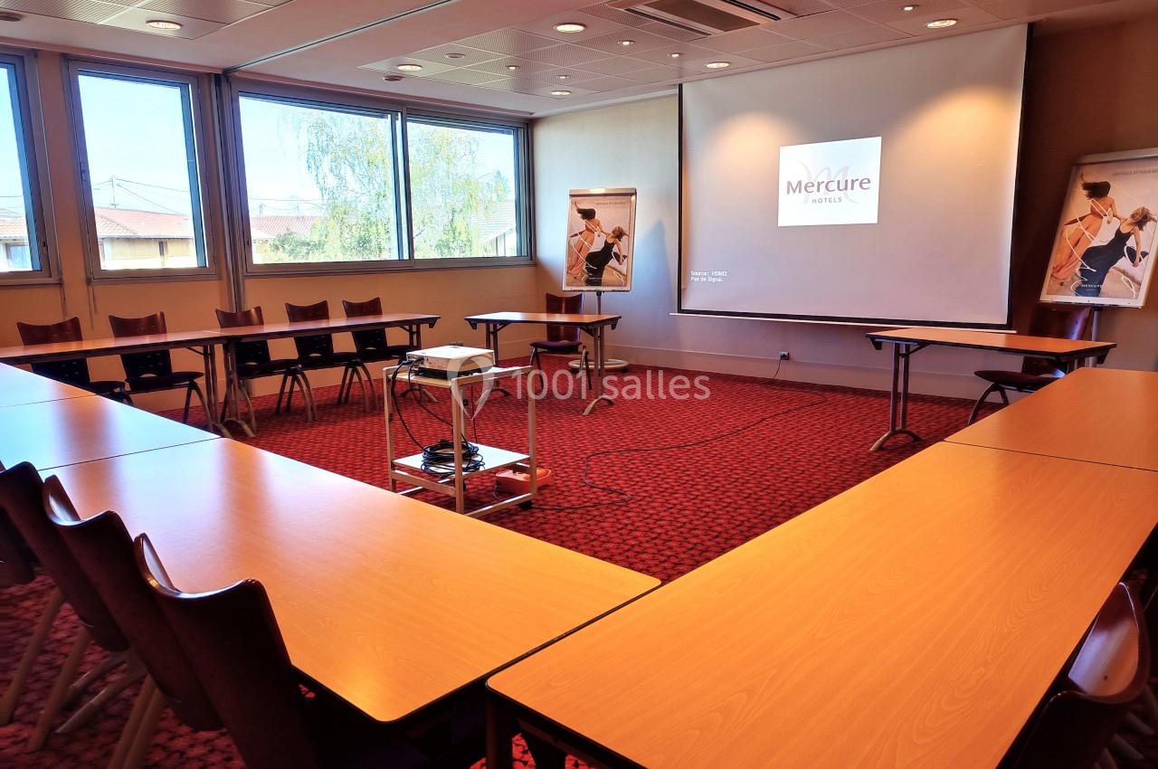 Salle de réunion lumineuse avec tables en U, projecteur allumé et affiches sur chevalets près des fenêtres.