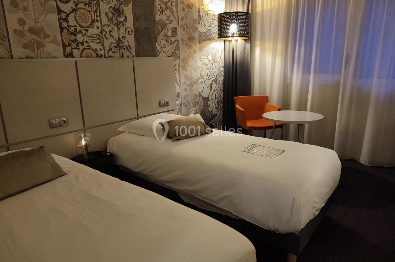 Deux lits simples dans une chambre d'hôtel avec décoration murale, lampe allumée, fauteuil orange et table ronde.