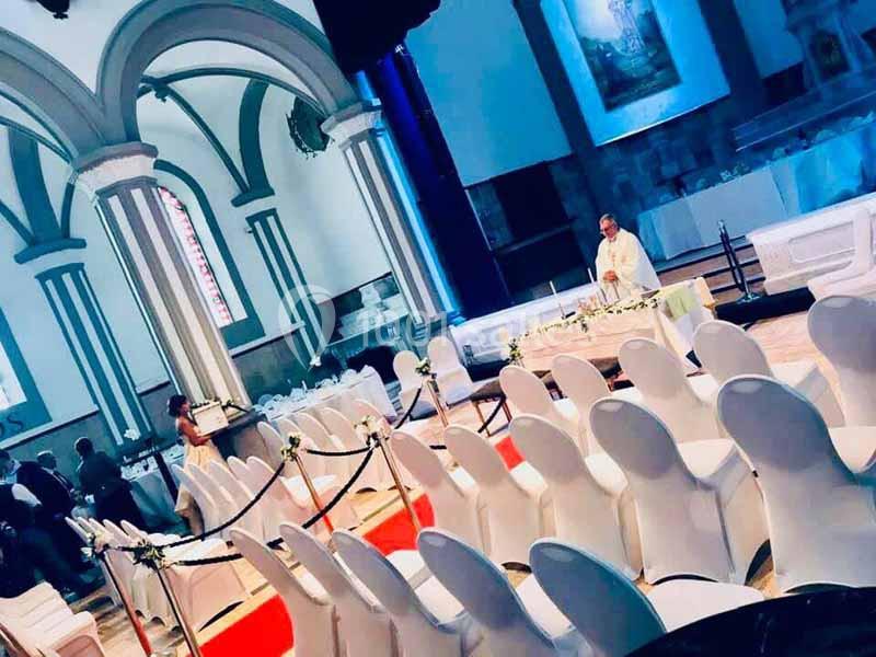 Salle de réception avec des chaises blanches alignées, un prêtre officiant près d'un autel et des décorations florales.