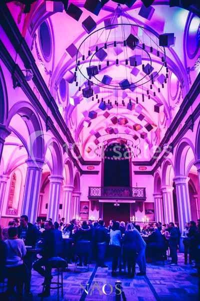 Intérieur d'une église transformée en lieu événementiel, éclairée par des lumières violettes, avec des tables et des…