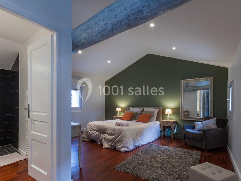 Chambre lumineuse avec lit double, mur vert, poutre apparente, parquet en bois et salle de bain attenante.