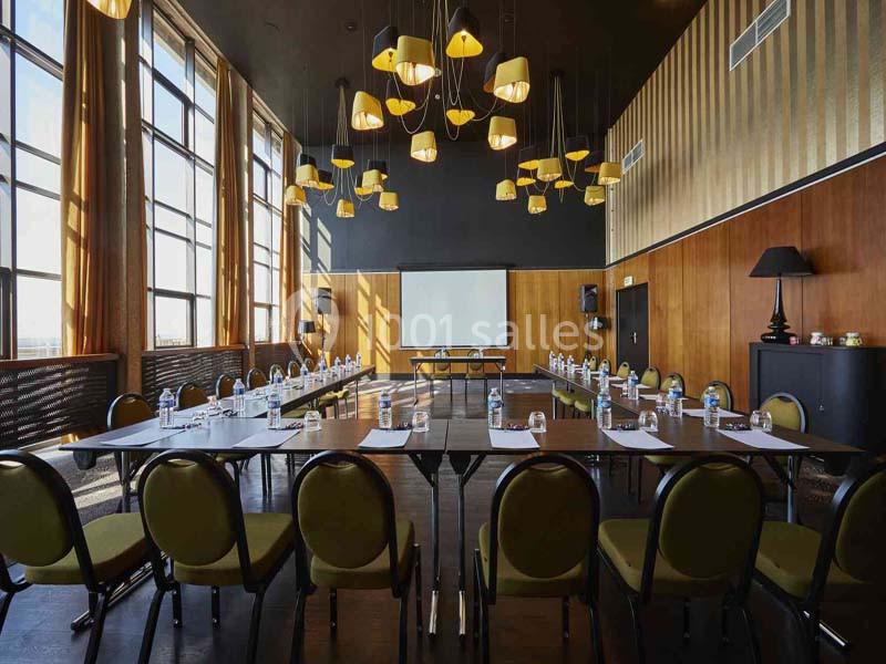 Salle de réunion lumineuse avec tables disposées en U, chaises vertes, écran de projection et lustres modernes.