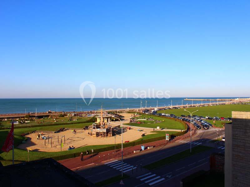Vue d'un parc avec une aire de jeux, une route, une pelouse et la mer à l'horizon sous un ciel dégagé.