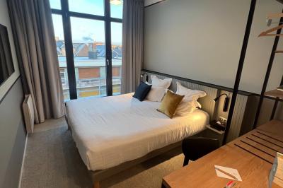 Chambre moderne avec lit double, bureau équipé, grande fenêtre avec vue sur les toits et éclairage suspendu.