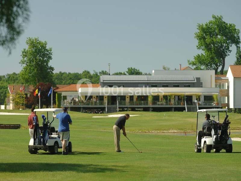 Un golfeur s'apprête à frapper une balle sur un terrain de golf, avec des voiturettes et un bâtiment en arrière-plan.