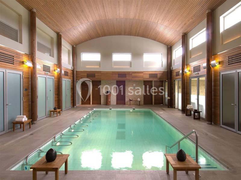 Piscine intérieure avec plafond en bois, entourée de cabines et de chaises en bois, dans un espace lumineux.