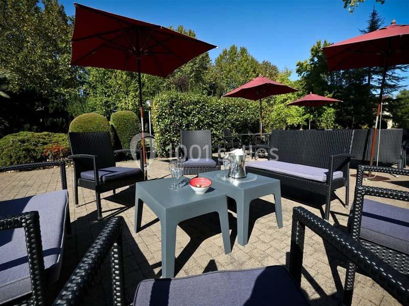 Mobilier de jardin en rotin noir avec tables grises, entouré de parasols rouges et de verdure sous un ciel bleu.