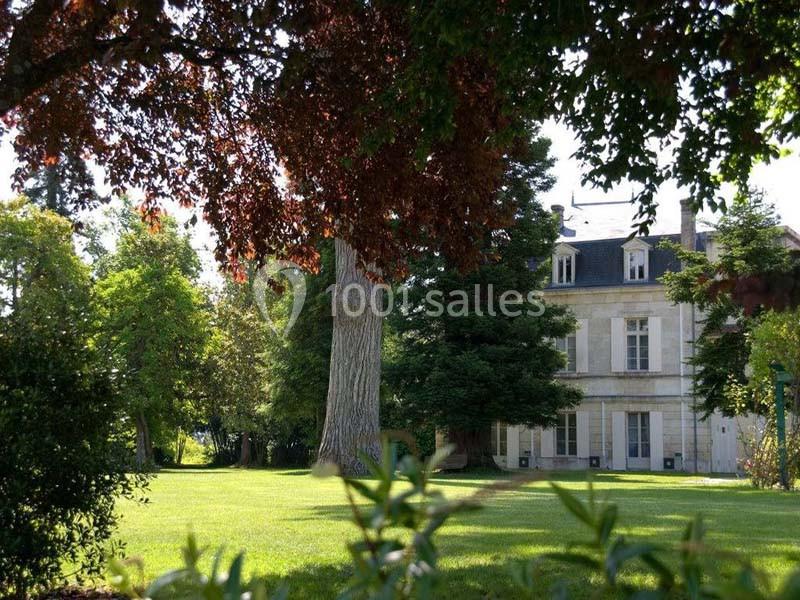 Vue d'un grand jardin arboré avec pelouse, encadrant une maison en pierre à deux étages sous un ciel dégagé.