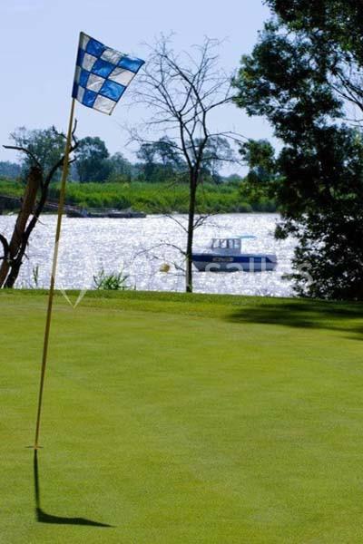 Drapeau de golf sur un green près d'une rivière, avec un bateau visible en arrière-plan et des arbres bordant l'eau.