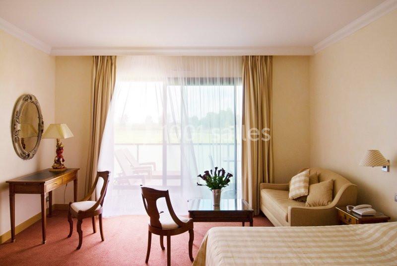 Chambre lumineuse avec lit, canapé, bureau, chaises, table basse et grande baie vitrée donnant sur un balcon.