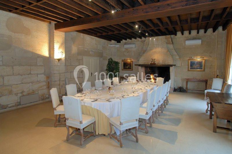 Salle de réception avec une grande table dressée, entourée de chaises blanches, dans un cadre en pierre avec cheminée.