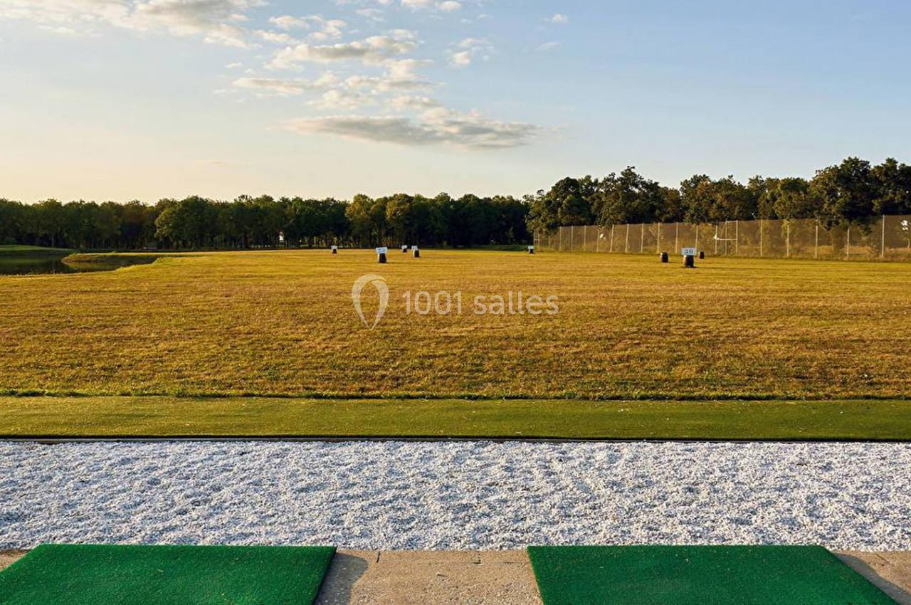 Terrain de golf avec zone d'entraînement en gazon, filets de protection et arbres en arrière-plan sous un ciel dégagé.
