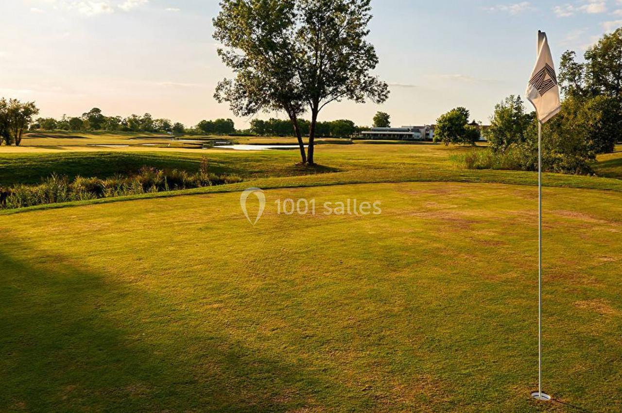 Vue d'un terrain de golf avec un drapeau sur le green, entouré d'arbres et d'une étendue d'herbe sous un ciel dégagé.