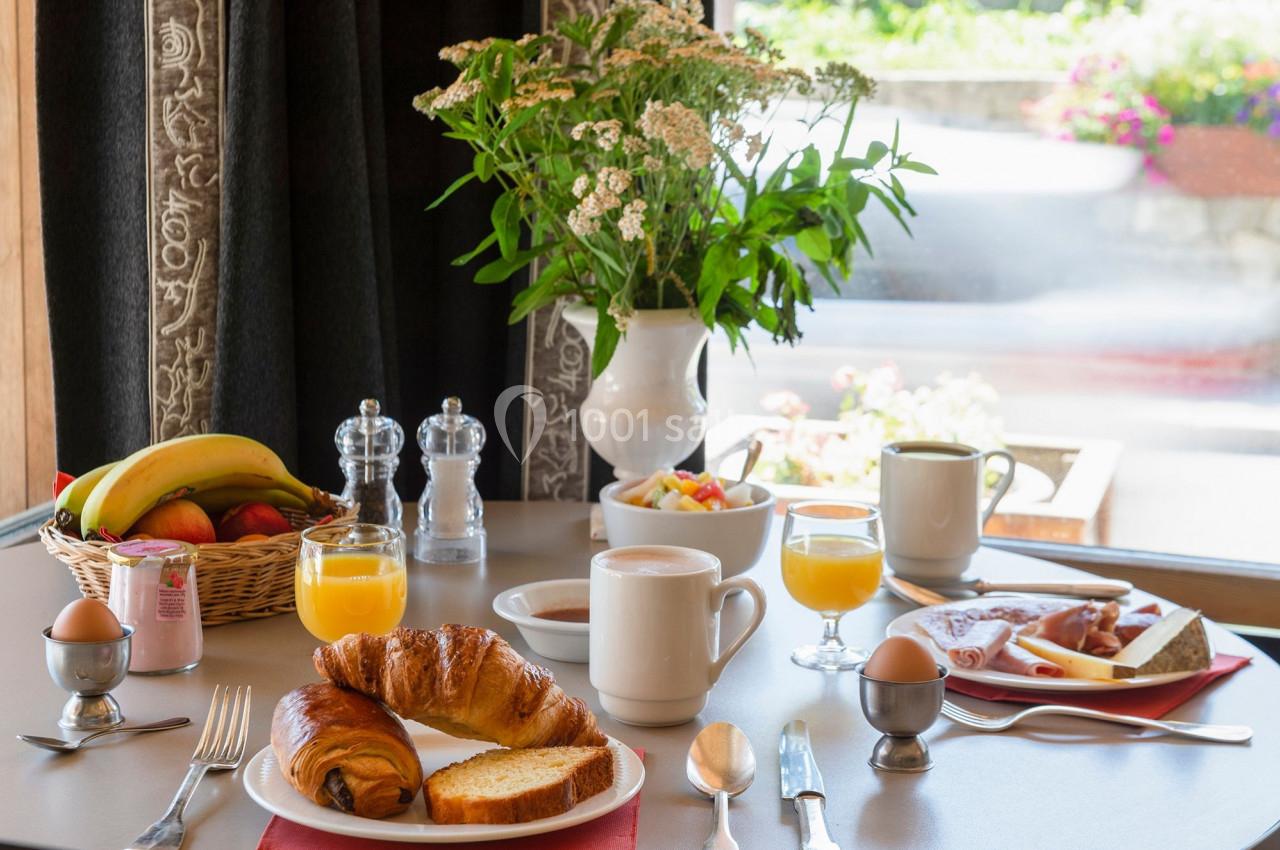 Petit-déjeuner composé de croissants, pain, œufs, fruits, jus d'orange, café et charcuterie sur une table près d'une fenêtre.
