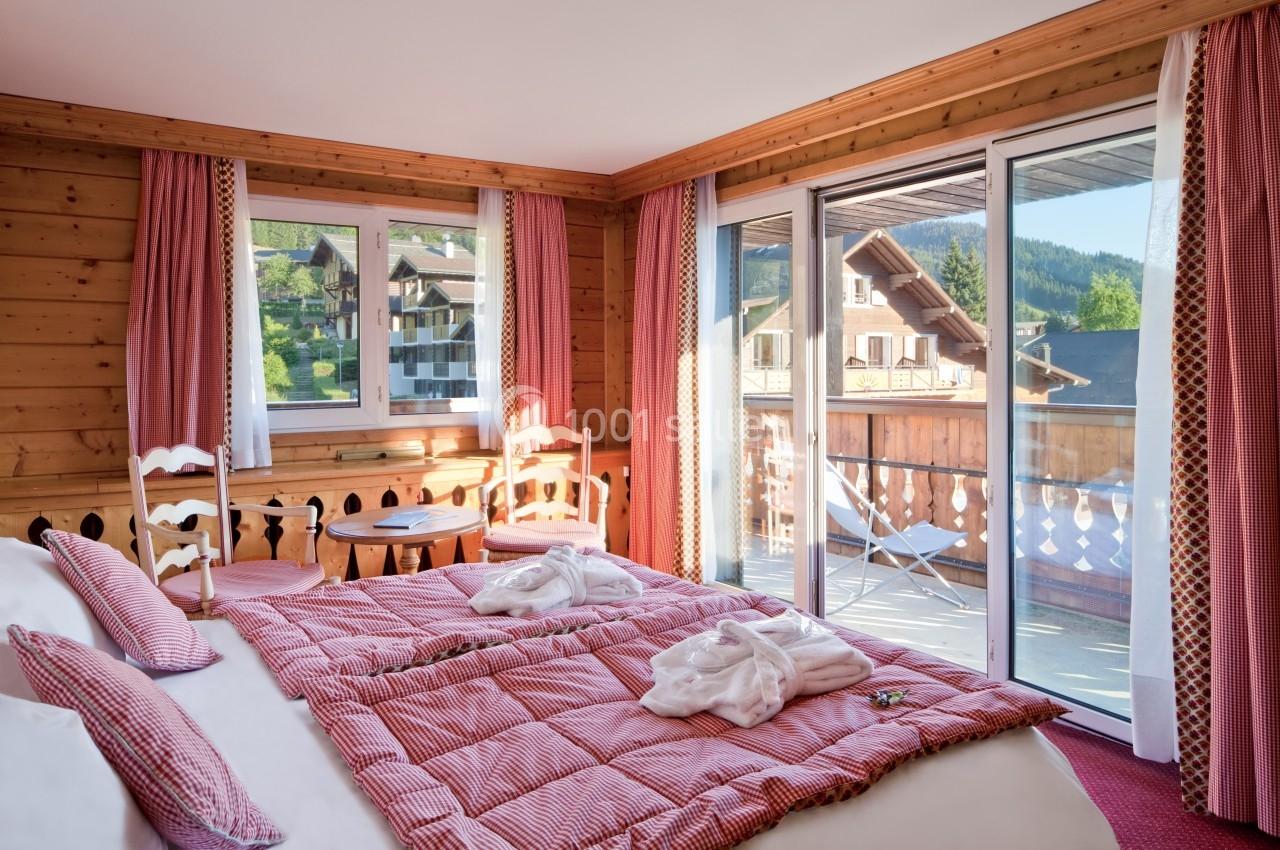 Chambre en bois avec lit double, balcon, grandes fenêtres et vue sur des chalets en montagne.