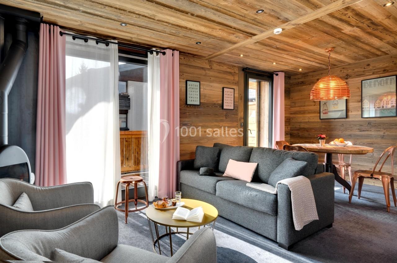 Salon chaleureux avec canapé gris, fauteuils, table basse, décoration en bois et lumière naturelle entrant par les fenêtres.