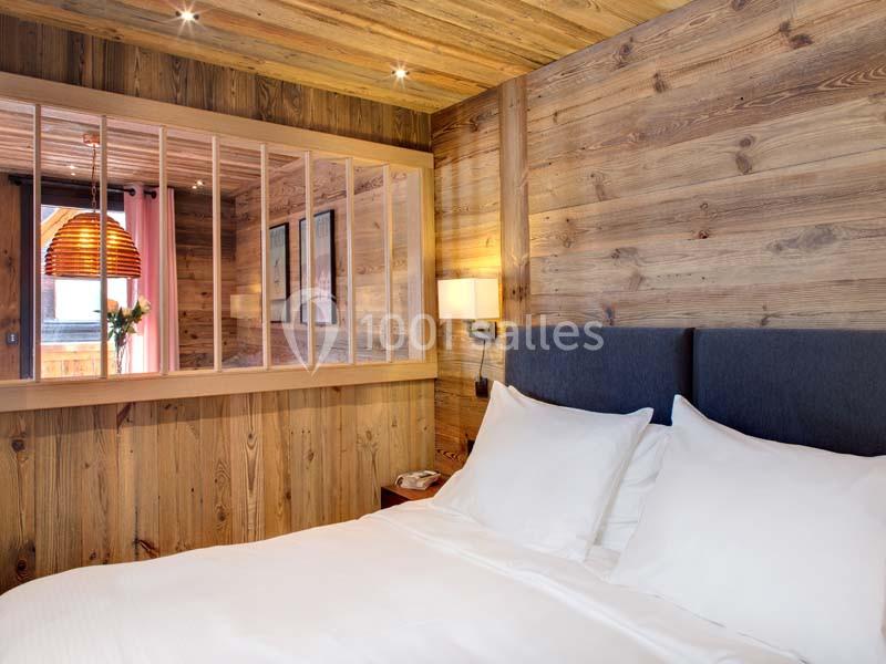 Chambre avec lit double, murs en bois, éclairage doux et cloison vitrée donnant sur un espace adjacent.