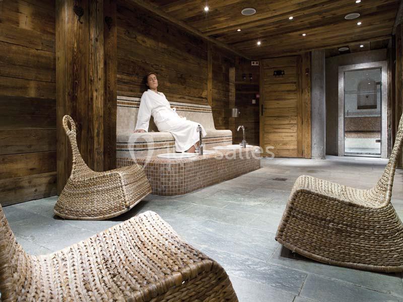 Une personne en peignoir se détend dans un spa en bois avec des fauteuils en rotin et un sol en pierre.