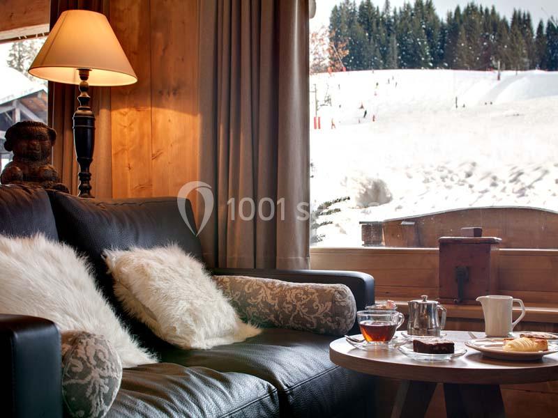 Salon cosy avec canapé, coussins, table basse avec thé et pâtisseries, vue sur un paysage enneigé par la fenêtre.