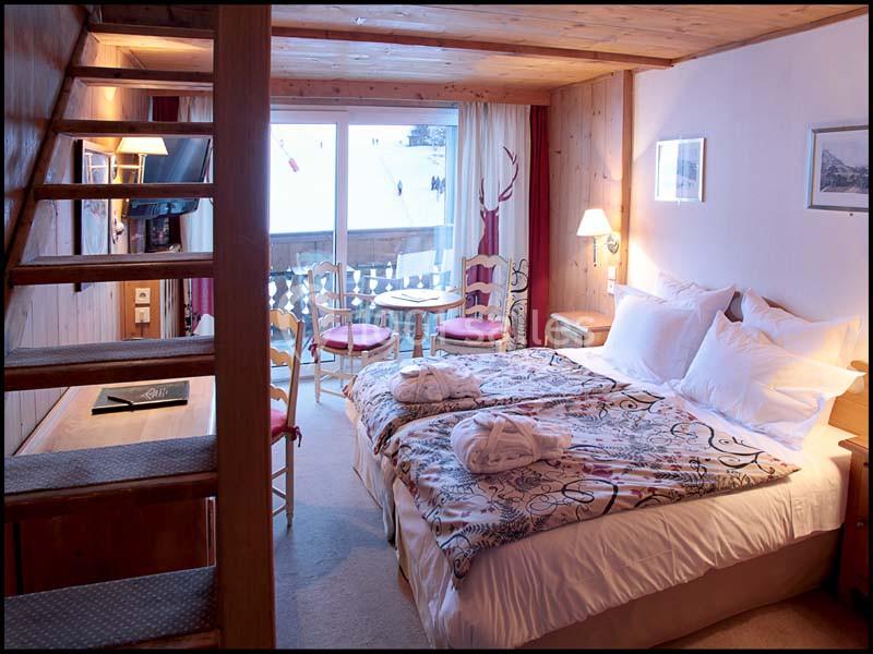 Chambre chaleureuse avec lit double, escalier en bois, balcon donnant sur un paysage enneigé et lumière naturelle.