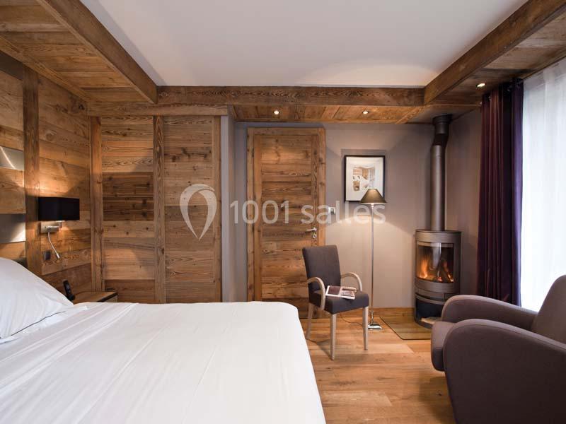 Chambre avec murs en bois, lit, fauteuils, poêle à bois allumé et lumière naturelle entrant par une grande fenêtre.