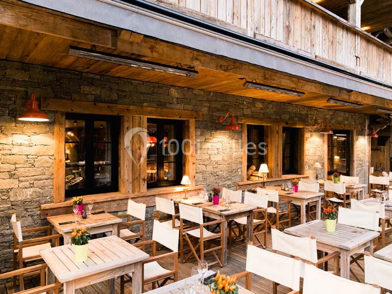 Terrasse en bois avec tables et chaises, décorée de plantes en pots, devant une façade en pierre et bois éclairée.