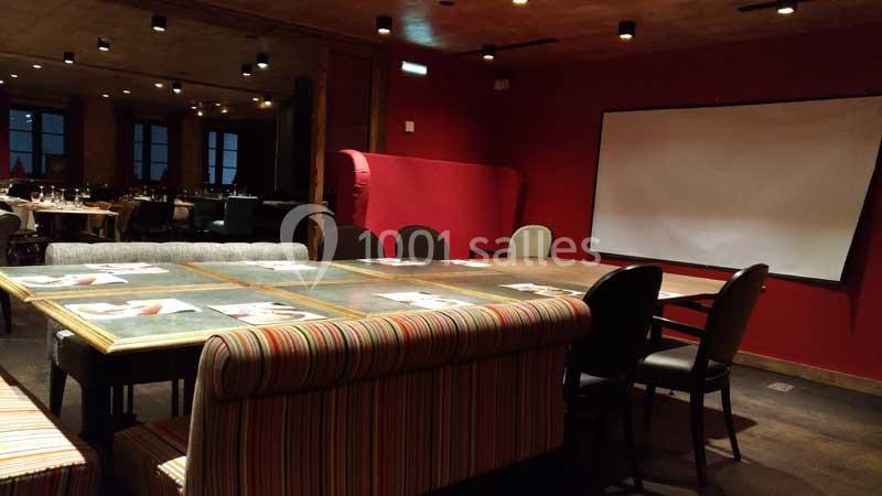 Salle de réunion avec grande table, chaises variées, banquette rayée et écran blanc, éclairage tamisé.