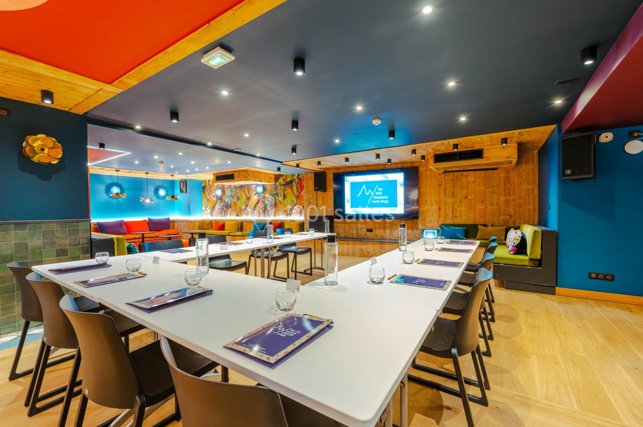 Salle de réunion colorée avec tables en U, chaises noires, écran de présentation et décorations murales vives.