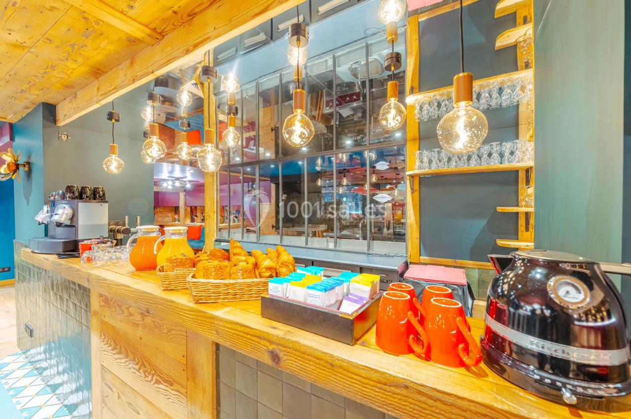 Buffet de petit-déjeuner avec jus, viennoiseries, tasses rouges et machine à café sur un comptoir en bois éclairé.