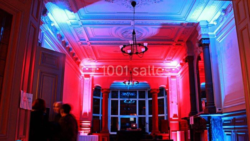 Salle avec un plafond orné, éclairée par des lumières rouges et bleues, avec des silhouettes floues au premier plan.